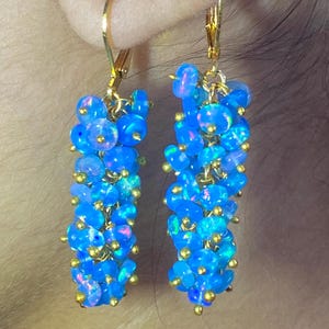 May include: A pair of dangle earrings featuring clusters of small, round, vibrant blue opals with iridescent flashes. The stones are attached to gold-toned metal wire and suspended from gold-toned lever-back hooks, creating a textured, elongated shape.