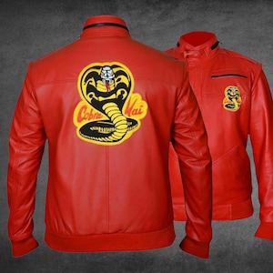 May include: A bright red jacket featuring a large yellow and black Cobra Kai snake emblem on the back and a smaller matching logo on the left chest. The jacket has a stand-up collar, long sleeves with ribbed cuffs, and a front zip closure.