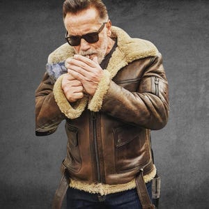 May include: A brown leather bomber jacket featuring a thick beige shearling lining, a wide collar, a front zip closure, and two large flap pockets on the chest. The jacket includes adjustable side straps at the hem and a small zipped pocket on the sleeve.