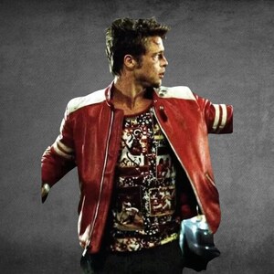 May include: A person with short dark hair wearing a red leather jacket with white shoulder and sleeve stripes over a patterned shirt featuring multiple rectangular graphic prints. The jacket is open, revealing the shirt underneath against a gray background.