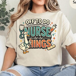 May include: A cream-colored t-shirt featuring a graphic of a goose wearing a nurse cap and stethoscope, holding a syringe. The text reads "OFF TO DO NURSE THINGS" in a retro font, accompanied by illustrations of a syringe, a bandage, and stars.