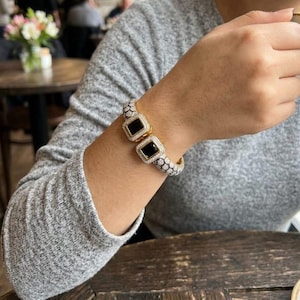 May include: A gold bracelet featuring two black square gemstones, each framed by white gemstones. The bracelet is worn on an arm, with a grey long-sleeved top visible. The design of the bracelet is detailed and elegant.