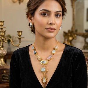 Può includere: Una collana color oro caratterizzata da una serie di frammenti di pietre preziose grezze e multicolori incastonati nel metallo, con un ciondolo centrale composto da due cornici dorate circolari e a goccia che racchiudono pietre.