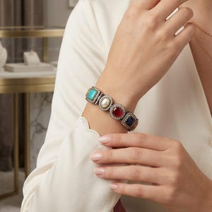 May include: A silver bracelet featuring five gemstone settings in square and rectangular shapes. The gemstones include turquoise, a pearl, ruby, and sapphire. The bracelet is worn on a wrist, with a white long-sleeved shirt.