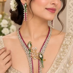 May include: A necklace and earring set featuring bird motifs with green and white stones, accented by rows of pink and clear crystals. The necklace includes a central branch design with two birds, while the earrings are long, dangling bird-shaped pieces.