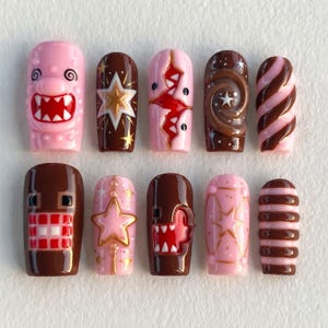 May include: A set of ten press-on nails featuring a variety of designs in pink and brown. The patterns include cartoon faces with sharp teeth, gold stars, swirling chocolate-like textures, and striped motifs, all finished with a glossy top coat.
