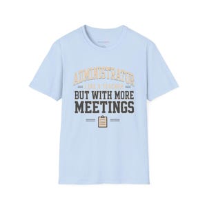 Op de afbeelding: Een lichtblauw t-shirt met korte mouwen voorzien van de tekst "ADMINISTRATOR LIKE A TEACHER BUT WITH MORE MEETINGS" in beige en donkergrijze letters, vergezeld van een kleine afbeelding van een beige klembord onder de tekst.