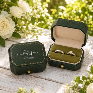May include: A dark green octagonal ring box with a gold-colored latch and trim. The lid features the white text h & j with a heart symbol and the date 10.12.2026. Inside, the box contains a silver-toned band and a gold-toned diamond ring.