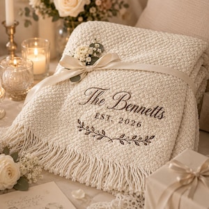 May include: A cream-coloured knitted throw blanket with fringe, featuring an embroidered design that reads The Bennetts EST. 2026 above a leafy branch motif. The blanket is wrapped with a smooth cream ribbon and adorned with a small floral sprig.