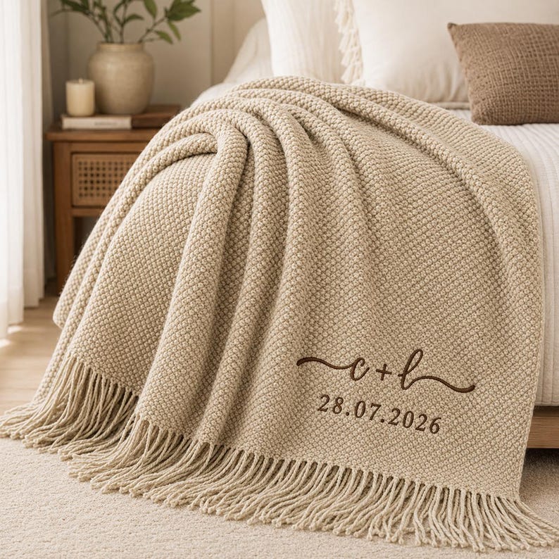 May include: A beige textured knit throw blanket with fringe trim. The blanket features dark brown embroidered text that reads c + l and the date 28.07.2026. The fabric has a consistent waffle-like pattern throughout the entire surface area.