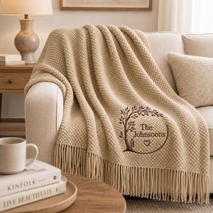 Op de afbeelding: Een beige, getextureerde gebreide plaid met franjes aan de randen, voorzien van een geborduurd ontwerp van een cirkelvormig takkenframe, een klein hartje en de tekst "The Johnsoons" in donkerbruine draad, gedrapeerd over een lichte bank.