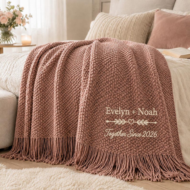 May include: A dusty rose textured knit throw blanket with fringe trim. The blanket features white embroidered text that reads Evelyn + Noah, a decorative arrow with a heart, and the phrase Together Since 2026, centered on the lower right side.