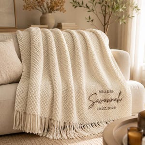 May include: A cream-colored knitted throw blanket with a textured bobble pattern and fringe trim. The blanket features dark brown embroidered text that reads MR. & MRS. Savannah 10.22.2026, positioned in the lower right corner of the fabric.