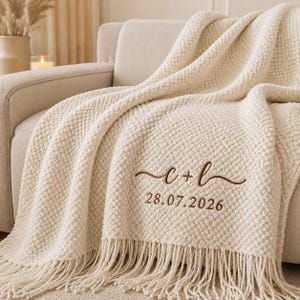 May include: A cream-colored textured knit throw blanket featuring a decorative fringe along the bottom edge. The blanket is embroidered with the brown text c + l in a cursive script, followed by the numerical date 28.07.2026 in a simple font.