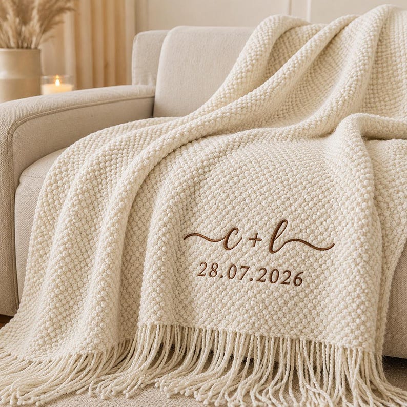 May include: A cream-colored textured knit throw blanket featuring a decorative fringe along the bottom edge. The blanket is embroidered with the brown text c + l in a cursive script, followed by the numerical date 28.07.2026 in a simple font.
