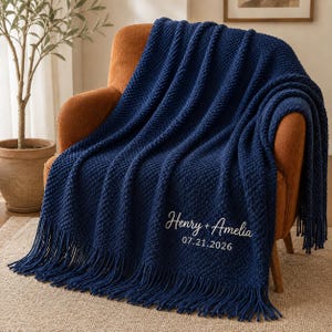 May include: A deep navy blue knitted throw blanket featuring a textured pattern and long fringe along the bottom edge. The blanket is embroidered with the white text Henry + Amelia and the date 07.21.2026 in a cursive and sans-serif font.