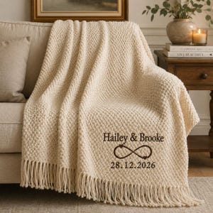 Op de afbeelding: Een crèmekleurige, getextureerde gebreide plaid met franjes aan de onderkant. De deken is geborduurd met de donkerbruine tekst Hailey & Brooke, een oneindigheidssymbool met twee kleine hartjes en de datum 28.12.2026.