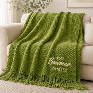 May include: A textured olive green knit throw blanket featuring a fringe edge and cream-colored embroidery that reads THE Bowman FAMILY. The blanket is draped over a neutral-toned sofa, showcasing a consistent popcorn stitch pattern throughout.