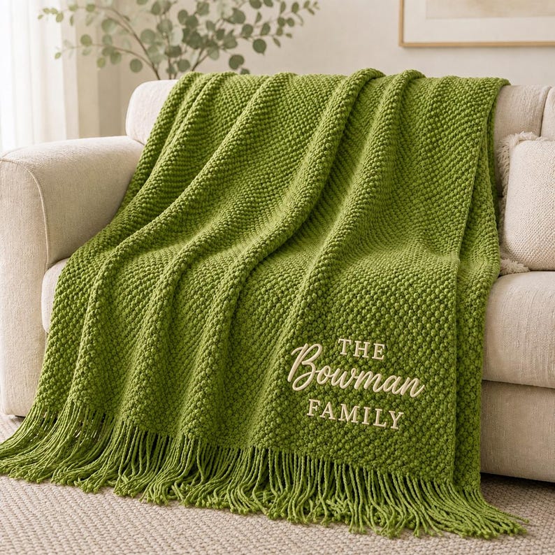 May include: A textured olive green knit throw blanket featuring a fringe edge and cream-colored embroidery that reads THE Bowman FAMILY. The blanket is draped over a neutral-toned sofa, showcasing a consistent popcorn stitch pattern throughout.