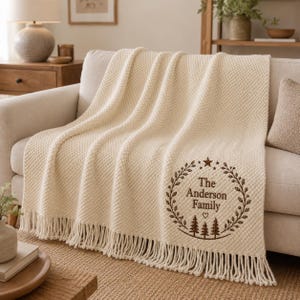 May include: A cream-coloured textured throw blanket with fringe edges. The blanket features an embroidered circular design in brown thread, including a star, a wreath of leaves, three pine trees, a heart, and the text The Anderson Family.