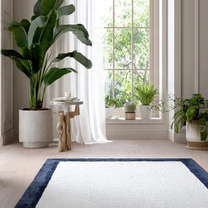 May include: A rectangular area rug featuring a solid white textured center panel framed by a thick, dark navy blue border. The rug lies flat on a light-colored wooden floor, showcasing a clean, simple design with a soft, plush pile construction.