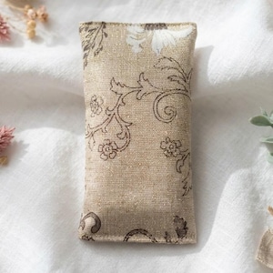 Könnte beinhalten: Ein rechteckiges Sachet aus einem weichen, strukturierten Stoff in einem hellen Beigeton. Die Oberfläche zeigt ein dunkelbraunes und weißes Blumenmuster mit geschwungenen Rankenmotiven und einem dezenten metallischen Schimmer.