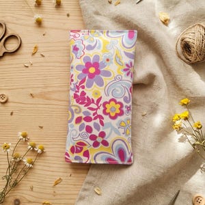Può includere: Una custodia rettangolare in tessuto con un vivace motivo floreale caratterizzato da fiori viola, magenta, gialli e azzurri su sfondo bianco. La custodia presenta una struttura piatta cucita e un design ripetuto di fiori e forme stilizzate.