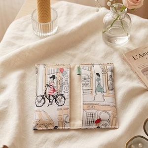 Pode incluir: Uma carteira de tecido bege aberta, com uma ilustração de uma cena de rua parisiense. A ilustração mostra uma mulher de bicicleta, edifícios e figuras. A carteira está sobre uma superfície bege, com uma vela e flores.