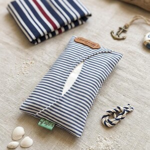 Pode incluir: Um estojo retangular de tecido para lenços com um padrão de riscas azul-marinho e branco. O estojo tem uma abertura central, uma etiqueta de couro castanho com a inscrição Hand made e uma pequena etiqueta verde com o texto lufex.