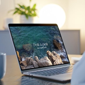 May include: A silver laptop displaying a scenic ocean view with the text "Petit à petit, Jour après jour" in white. The laptop is open on a wooden surface, with a blurred background.