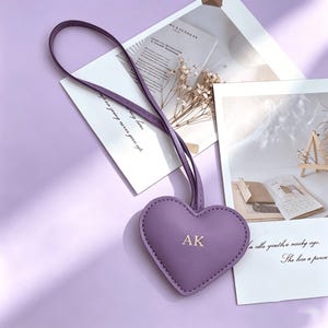 Könnte beinhalten: Ein herzförmiger Schlüsselanhänger in Lavendel mit den Initialen "AK" in goldfarbenen Buchstaben. Der Schlüsselanhänger hat einen passenden Lavendelriemen. Der Hintergrund umfasst weiße Karten mit Text und Trockenblumen, alles auf einer Lavendeloberfläche.