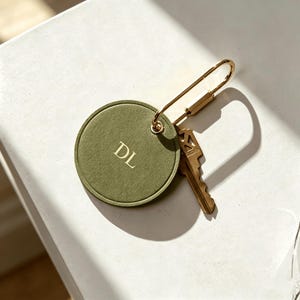 Könnte beinhalten: Olivgrüner Schlüsselanhänger mit goldfarbener Hardware und einem Schlüssel. Der Anhänger ist rund und mit den Initialen "DL" in Gold geprägt. Der Schlüsselring ist an einem goldfarbenen Sicherheitsnadel-Verschluss befestigt.