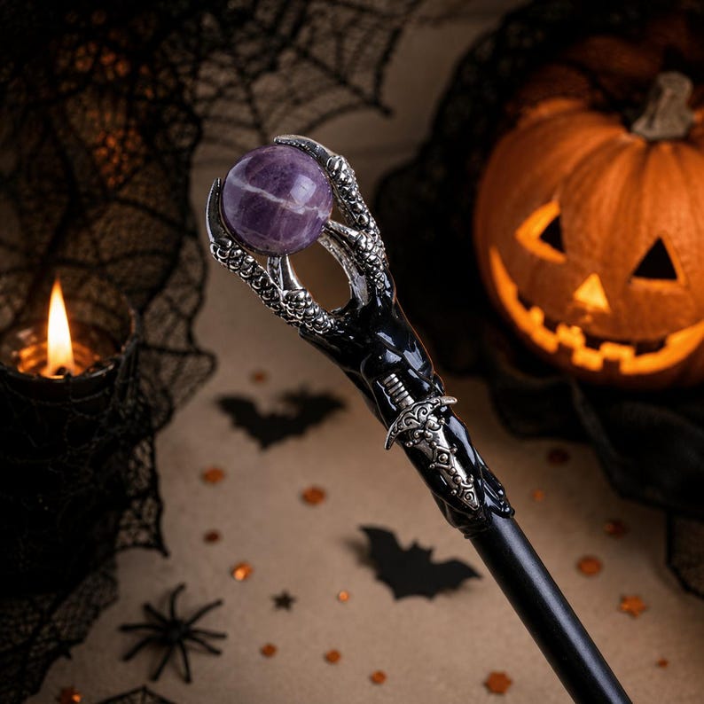 May include: A black wand featuring a silver-toned claw at the top holding a round, purple-striped stone sphere. The handle is decorated with a small, intricate silver-toned dagger emblem integrated into the smooth, dark, cylindrical shaft.
