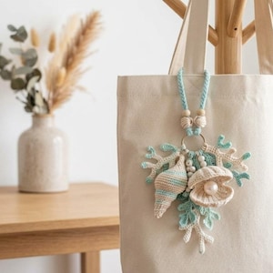 Può includere: Una borsa tote in tela beige con un ciondolo all'uncinetto appeso. Il ciondolo è composto da un cordino intrecciato azzurro, perline di legno ed elementi marini all'uncinetto, tra cui una conchiglia a spirale, un'ostrica aperta con perla e coralli.