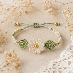 May include: A macrame bracelet featuring a central white flower with a gold bead center, flanked by two green leaf-shaped woven elements. The band is made of thin green cord with small gold beads and an adjustable sliding knot closure mechanism.