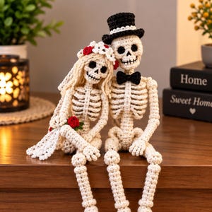 May include: Two crocheted skeleton figures sitting side by side. The figure on the left wears a white floral crown with red accents and a white veil. The figure on the right wears a black top hat with a white band and a black bow tie.