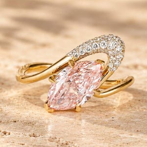 Op de afbeelding: Een gouden ring met een centrale lichtroze edelsteen in marquise-slijpvorm. De ringband buigt om de steen heen, waarbij één zijde is geaccentueerd met een groep kleine, heldere, rond geslepen steentjes langs het bovenste deel van de zetting.