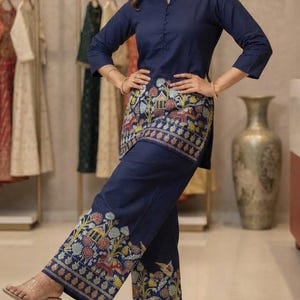 May include: A navy blue two-piece set consisting of a three-quarter sleeve tunic and wide-leg trousers. Both pieces feature a colorful, intricate folk-art style print along the hemline, depicting stylized elephants, trees, and floral patterns.