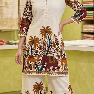 May include: A cream-colored tunic and wide-leg pants set featuring a repeating print of brown palm trees, dark red elephants, and camels. The tunic has three-quarter sleeves, a V-neckline with a button placket, and brown leather sandals.