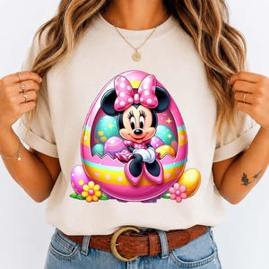 May include: A beige t-shirt featuring a colorful graphic of Minnie Mouse inside a pink Easter egg. The egg is decorated with pastel colors and surrounded by Easter eggs and flowers. Minnie Mouse is wearing a pink dress and bow.