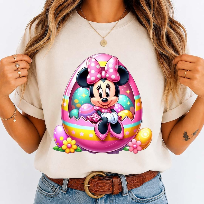 May include: A beige t-shirt featuring a colorful graphic of Minnie Mouse inside a pink Easter egg. The egg is decorated with pastel colors and surrounded by Easter eggs and flowers. Minnie Mouse is wearing a pink dress and bow.