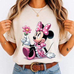 May include: Beige t-shirt featuring a cartoon Minnie Mouse in a pink polka-dot dress and bow, holding a pink flower. The design is centered on the shirt, with the character sitting. The shirt has short sleeves and a round neckline.