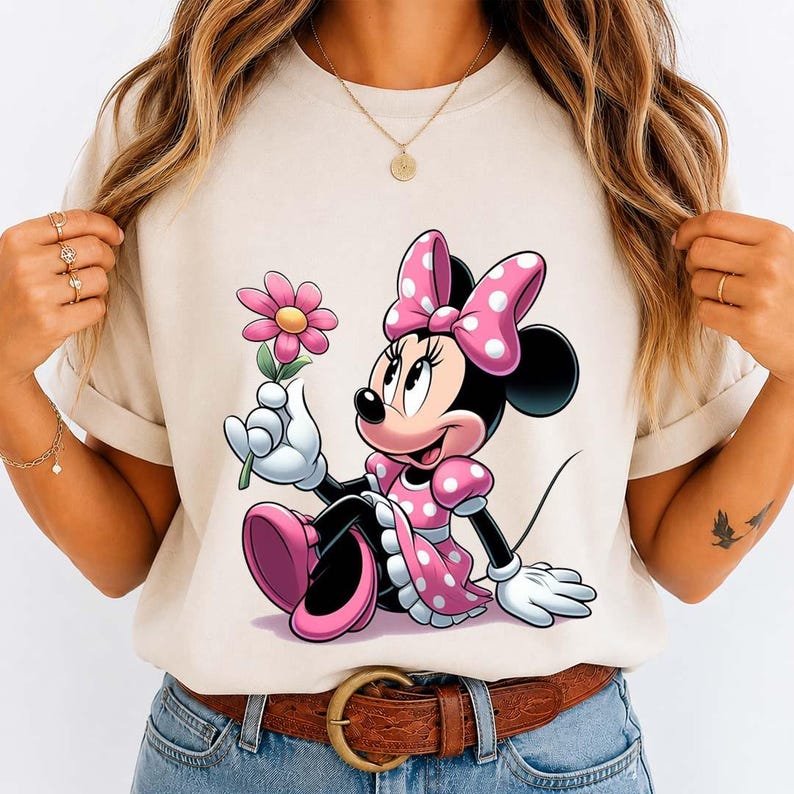 May include: Beige t-shirt featuring a cartoon Minnie Mouse in a pink polka-dot dress and bow, holding a pink flower. The design is centered on the shirt, with the character sitting. The shirt has short sleeves and a round neckline.