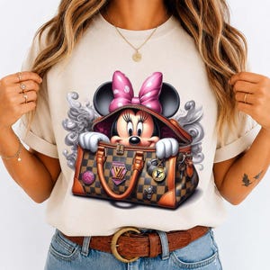 May include: Beige t-shirt featuring a Minnie Mouse graphic. Minnie is peeking out of a brown and tan checkered handbag with a pink bow. The handbag has gold hardware and a pink and gold logo. The graphic is surrounded by a gray swirl design.