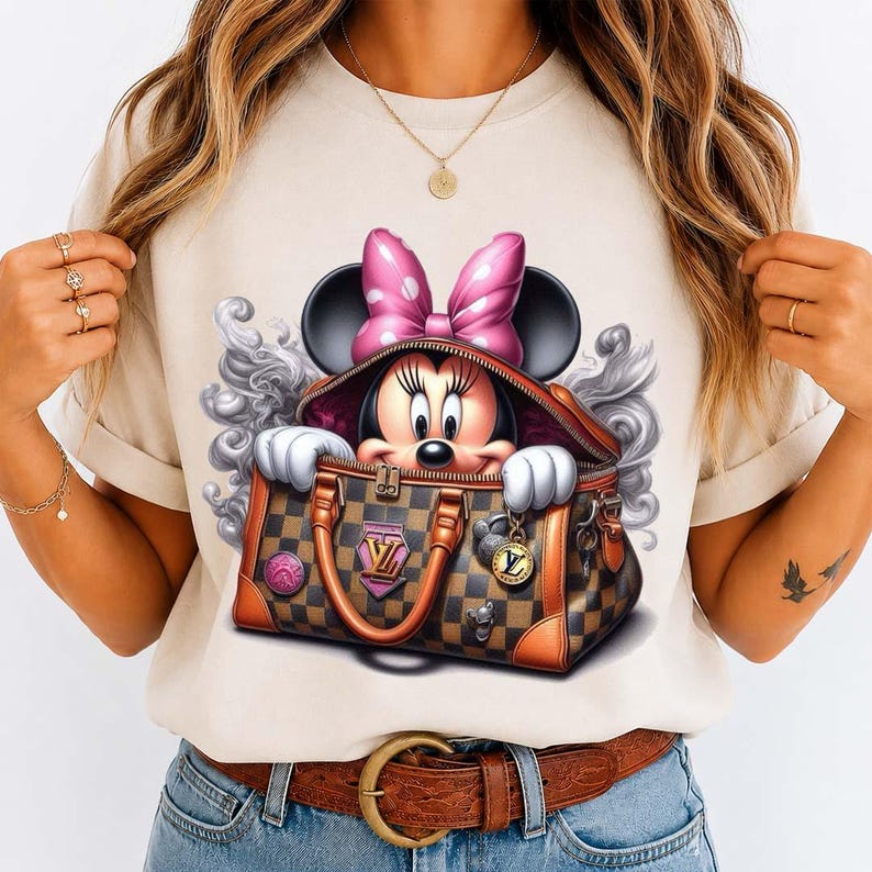 May include: Beige t-shirt featuring a Minnie Mouse graphic. Minnie is peeking out of a brown and tan checkered handbag with a pink bow. The handbag has gold hardware and a pink and gold logo. The graphic is surrounded by a gray swirl design.
