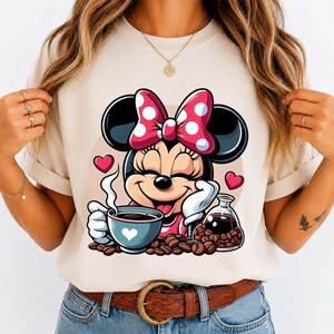 May include: A beige t-shirt featuring a cartoon Minnie Mouse with a red and white polka dot bow, holding a coffee cup with a heart design. Coffee beans and a glass carafe are also present.