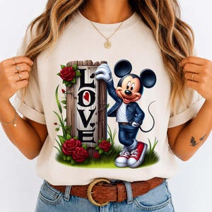 May include: A beige t-shirt featuring a graphic of Mickey Mouse in a denim jacket and jeans, leaning on a wooden sign that spells "LOVE" with a heart. Red roses and green foliage surround the sign and Mickey.
