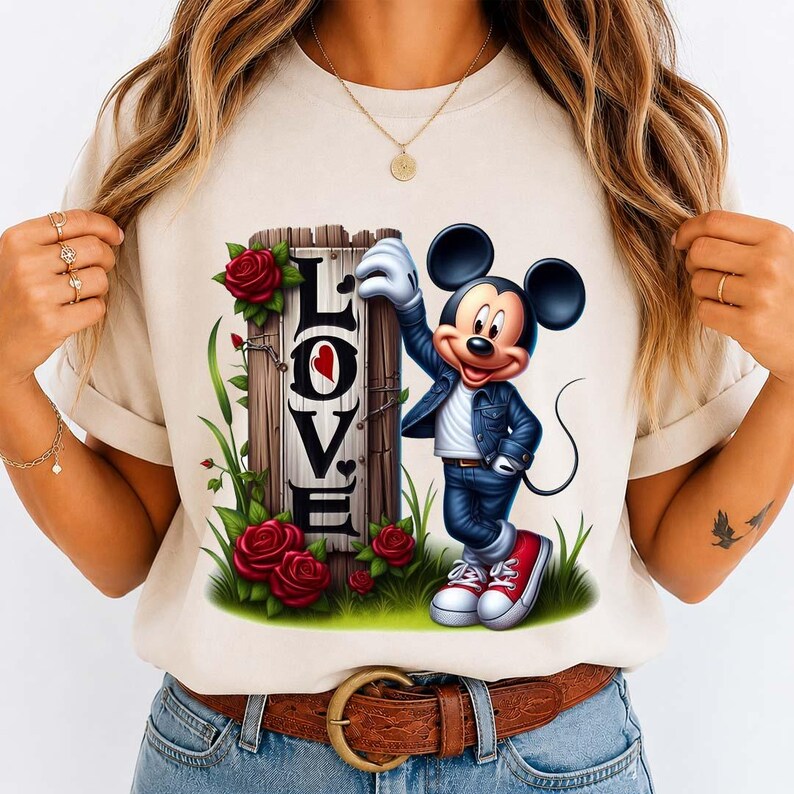 May include: A beige t-shirt featuring a graphic of Mickey Mouse in a denim jacket and jeans, leaning on a wooden sign that spells "LOVE" with a heart. Red roses and green foliage surround the sign and Mickey.