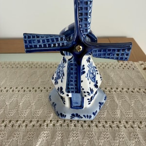 Op de afbeelding: Een keramische windmolenfiguur in blauw en wit. De windmolen heeft gedetailleerde blauwe wieken en een decoratief lichaam met bloemmotieven. De basis is zeshoekig en het ontwerp doet denken aan Delfts blauw.