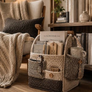 May include: A multi-compartment crochet organizer basket in shades of cream, beige, and dark brown. The basket features exterior pockets holding a pair of glasses and a ceramic mug, with a main section containing books and notebooks.